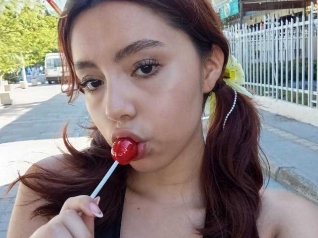 LolaBubbles with brown hair in pigtails, holding a red lollipop in her mouth, sucking it gently. She's wearing a black top and a yellow hair accessory.