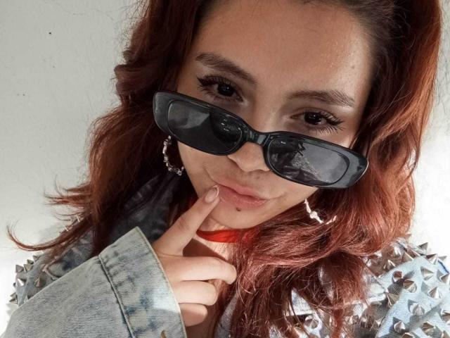 LolaBubbles with red wavy hair, wearing black sunglasses, large hoop earrings, and a studded denim jacket, has her finger on her pouty lips, looking sexy.