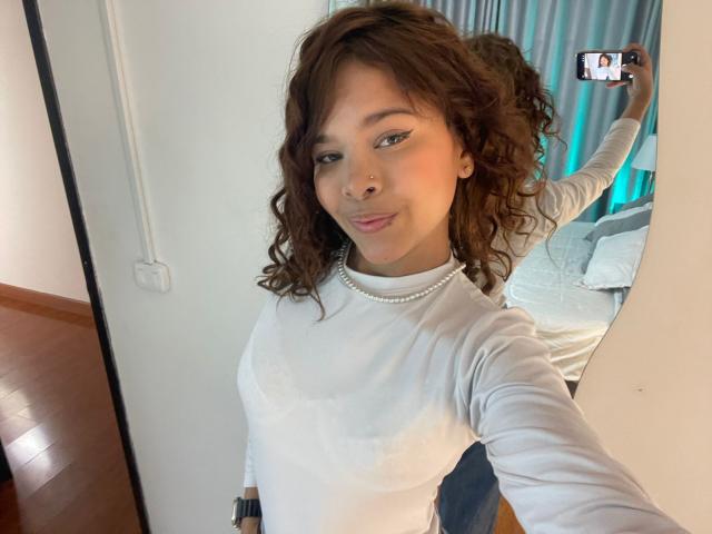 ChloeVixen, con i capelli castano ricci, indossa una camicia bianca a maniche lunghe, una collana di perle e rossetto rosa, mentre si scatta un selfie, mostrando il suo seno piccolo e i glutei nello specchio..