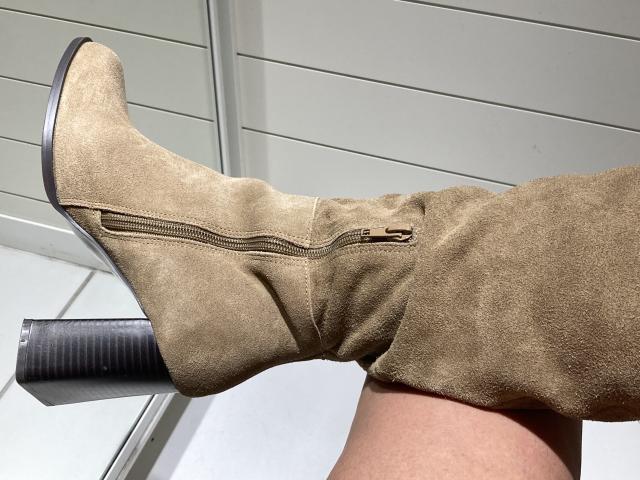 DeesseBleue's tan suede boot with a black heel and gold zipper, resting on a white surface, showing her tan leg and slight boot crease.