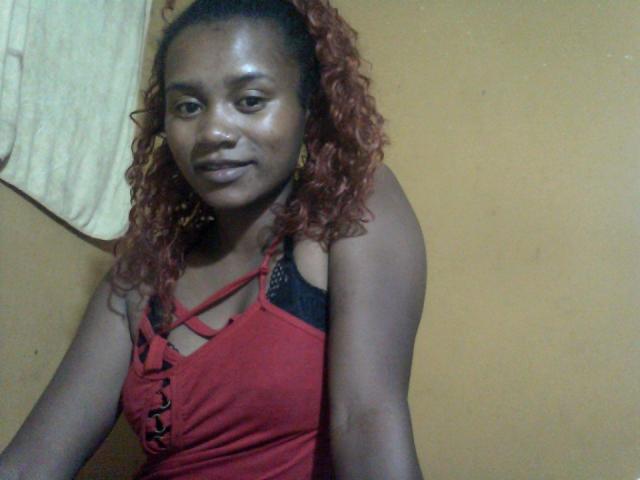 SousouHot with dark brown skin, curly red hair, wearing a red tank top showing black bra straps, and gold earrings, sitting and smiling softly.