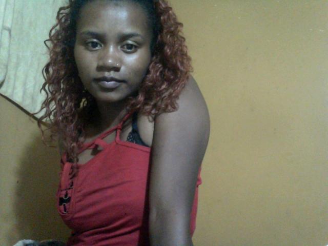 SousouHot has dark skin, curly red hair, and wears a red tank top with black straps, showing a small black bra. Her nose is pierced. She looks at the camera.