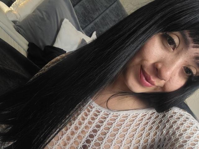 LunaBellaHotty with long black hair, smiling, wearing a white fishnet top, showing her tits through the fabric, in a bedroom. Her brown eyes and pink lips are visible.