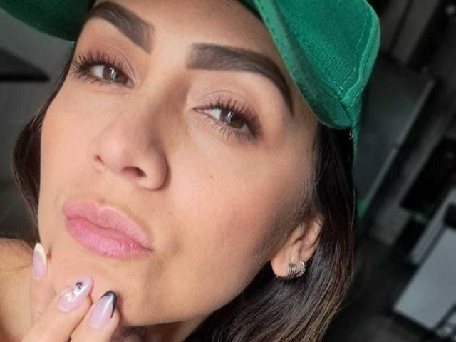 AriaRous has pouty pink lips, dark eyebrows, green cap, brown eyes, and is biting her finger with black and white nails. She's wearing a diamond earring.