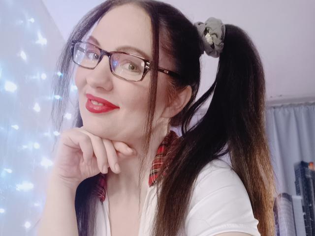 DashaHart with pigtails, red lipstick, glasses, and white shirt, resting chin on hand, smiling, red plaid collar, gray scrunchie, fair skin, red lips, brown hair, soft light.