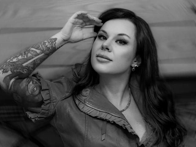 MissTwinkleX with tattoos on her arm, wearing a ruffled shirt, looking up with a slight smile, hand in her hair, black and white, earrings, and a necklace.