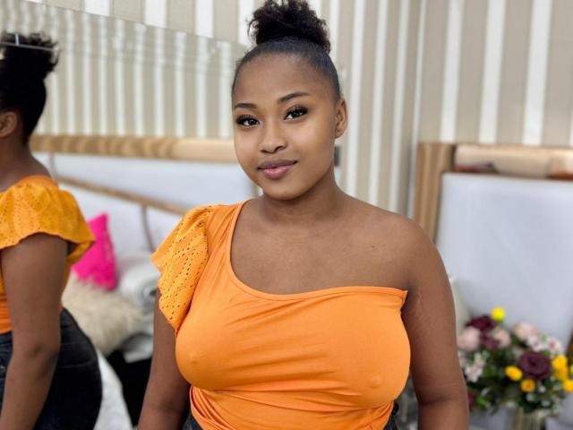 RailyBenet with medium brown skin, black hair in a bun, orange off-shoulder top, showing her big tits, standing in front of a mirror, smiling subtly.