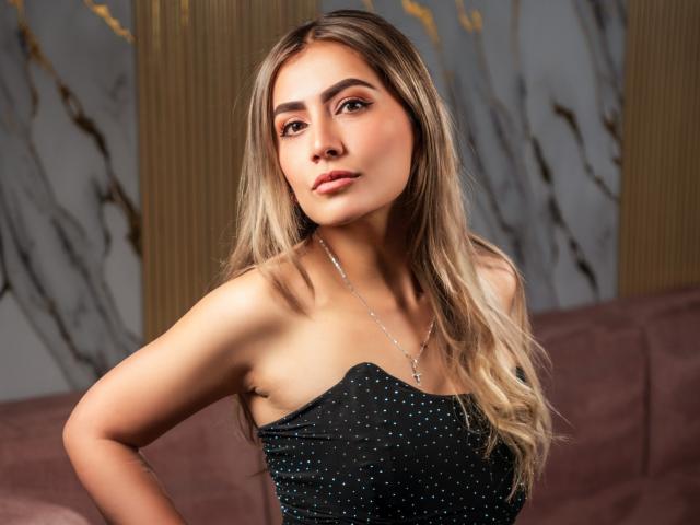 AriaRous with long blonde hair, wearing a black strapless dress with small blue dots, a silver necklace, and a confident expression, her tits slightly visible.