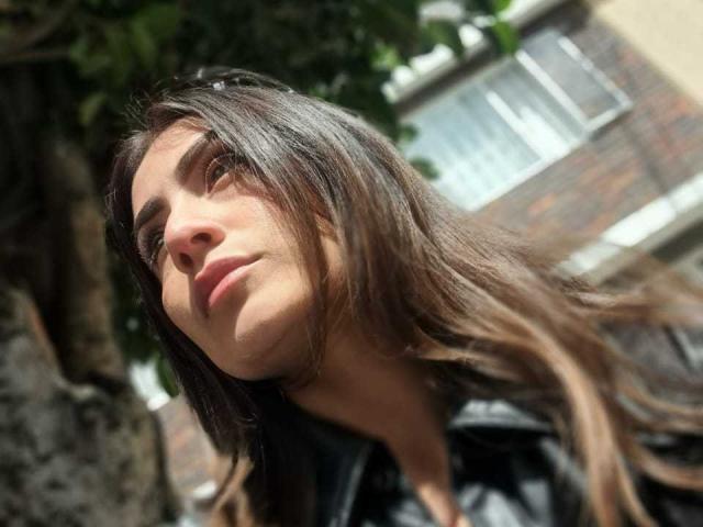 AriaRous has long brown hair, slightly wavy, with a small nose and light makeup. She's looking up with a subtle smile, wearing a black shirt. She's in sunlight.