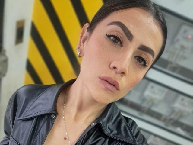 AriaRous with pouty lips, dark eyebrows, eyeliner, and a black leather jacket, wearing a necklace, has brown hair pulled back and small earrings.