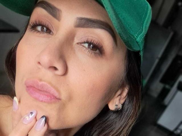 AriaRous with a green cap, brown eyes, thick eyebrows, pink lips, and manicured nails touching her chin. She's wearing a diamond earring. Her face is centered.