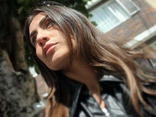 AriaRous has long brown hair, light skin, and brown eyes, wearing a black leather jacket, looking up with a slight smile, sunlight in her face, outdoors.
