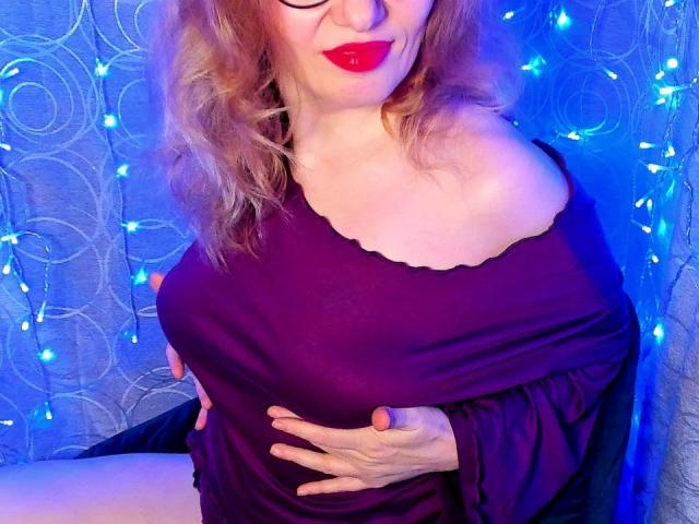 PaulinaSunny with red lipstick, wavy blonde hair, and off-shoulder purple top, holding her big tits, blue fairy lights in background.