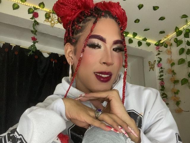 FlorenceLambert with red braided buns, purple eyeshadow, dark lipstick, white hoodie, finger ring, and purple nail polish, smiling, hands near face, with vines in the background.