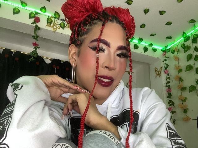 FlorenceLambert has red braided puffs, dark purple lipstick, winged eyeliner, white hoodie, and green LED strip lights in the background. She's posing with a finger near her mouth.