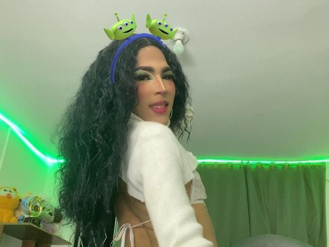 FlorenceLambert with curly black hair, green eyeshadow, white crop top, green headband with alien emojis, smiling, showing her back, green neon light in background.