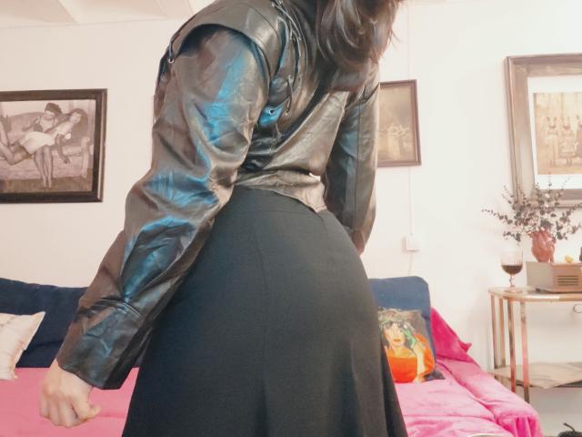 Chili está curvada, vestindo uma jaqueta de couro preta e uma saia preta apertada, expondo sua bunda curvada, em um quarto.