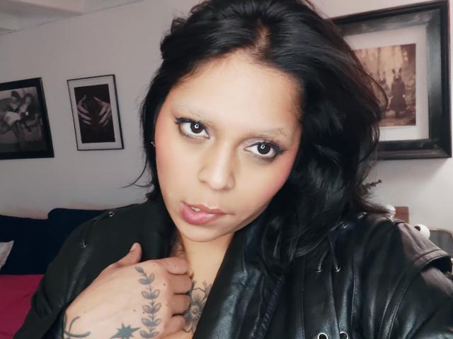 Chili con el pelo negro, delineador de ojos pesado y lápiz labial rosado mira seductoramente mientras toca su cuello tatuado con una mano, usando una chaqueta de cuero negra.