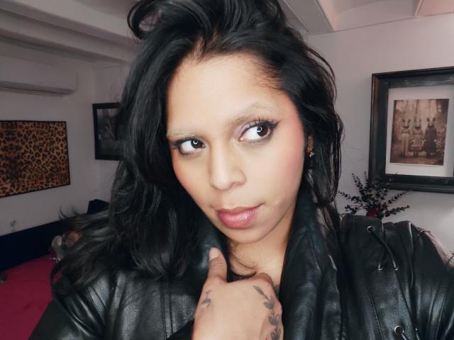 Chili, een sexy solo milf met zwart haar en zware make-up, kijkt recht in de camera, haar hand op haar leren jas.