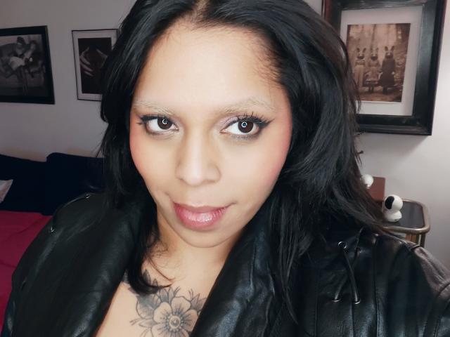 Chili tiene un tatuaje floral en el pecho, cabello negro y labios rosados brillantes, con ojos marrones hiper-sexy, mirando directamente a la cámara. ahead.