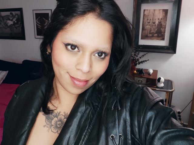 Chili, una donna sexy dalla pelle marrone con i capelli neri, giacca di pelle e tatuaggio floreale, sorride alla fotocamera, indossando rossetto rosa lucido.