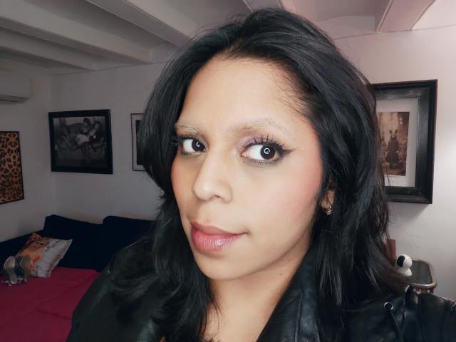 Chili, een sexy bruine vrouw met zwart haar en roze lippenstift, staart naar de camera met een brede, paarse blik.