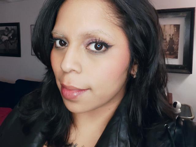 Chili avec des cheveux foncés et des lèvres roses brillantes, portant du cuir noir, regardant la caméra avec des yeux bruns étincelants, léger sourire.