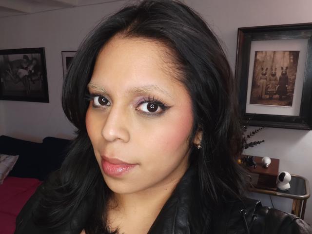 Chili tiene el pelo oscuro y brillante maquillaje, mirando directamente con grandes ojos marrones, vistiendo una chaqueta negra, mirando sexy como mierda..