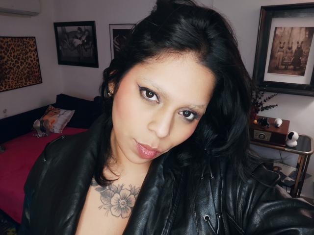 Chili, eine MILF mit schwarzen Haaren, schwerem Make-up und einem Blumen-Tattoo, starrt verführerisch in die Kamera und trägt einen schwarzen jacke aus Leder.