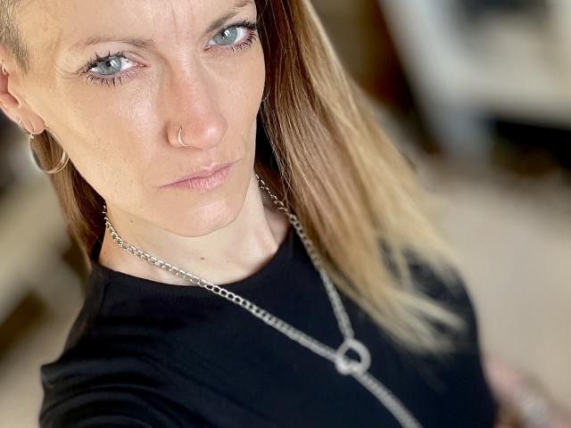 DaneeSoHot a un anneau au nez, des yeux bleus et des cheveux blonds, et elle porte une chemise noire avec un collier en argent. Elle regarde l'appareil photo avec une expression sérieuse..