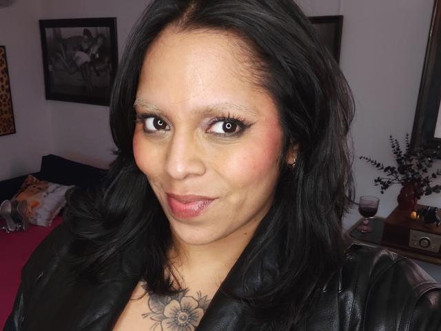 Chili hat schwarze Haare, rosa Lippen und ein Blumen-Tattoo auf ihrer Brust, während sie eine schwarze Lederjacke trägt..