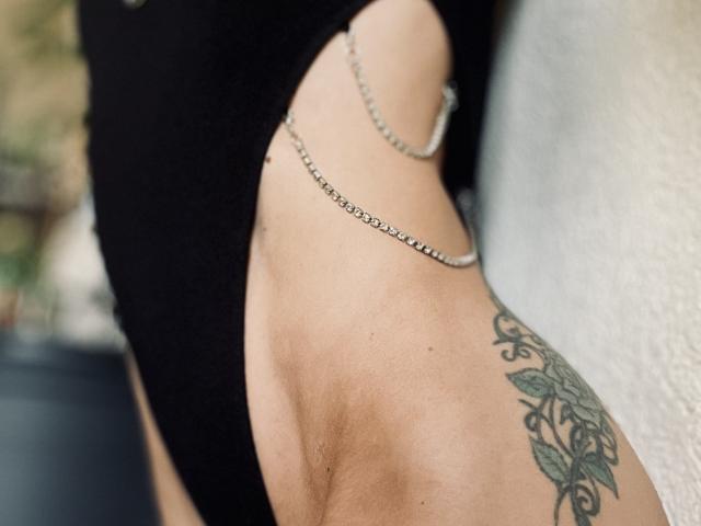 DaneeSoHot's side with a black one-piece, chain hanging from her hip, and a detailed tattoo of green leaves on her right hip.