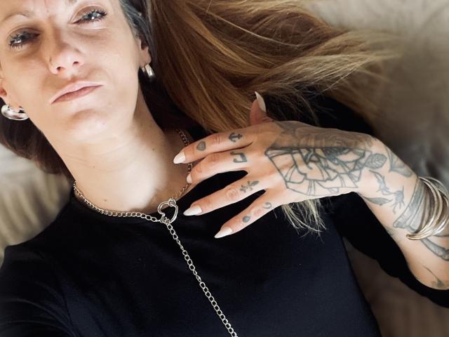 DaneeSoHot, con tatuaggi sulla mano, indossa una maglietta nera, una collana a catena e orecchini d'argento, è sdraiata, con una mano sul petto, e guarda verso la fotocamera..