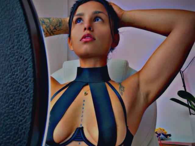 Loren, con la pelle abbronzata, i capelli neri raccolti in uno chignon, indossa un top nero con spalline sottili che mette in mostra il seno e ha un tatuaggio sotto il petto, con le braccia alzate e lo sguardo rivolto verso l'alto..
