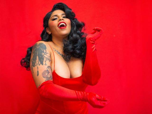 Chili está recibiendo un creampie mientras lleva guantes rojos y un vestido rojo escotado, con un brazo tatuado y cabello negro.