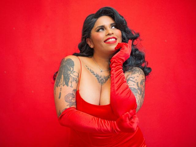 Chili con tatuaggi completi indossa un abito rosso scollato e lunghi guanti rossi, sorridente con il rossetto rosso, toccandola guancia.