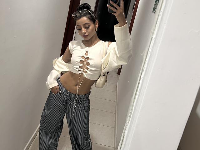 ThaliaCollins has small tits, wearing a white lace-up crop top, grey high-waisted jeans, and a white sweater pulled down, standing in a mirror selfie.