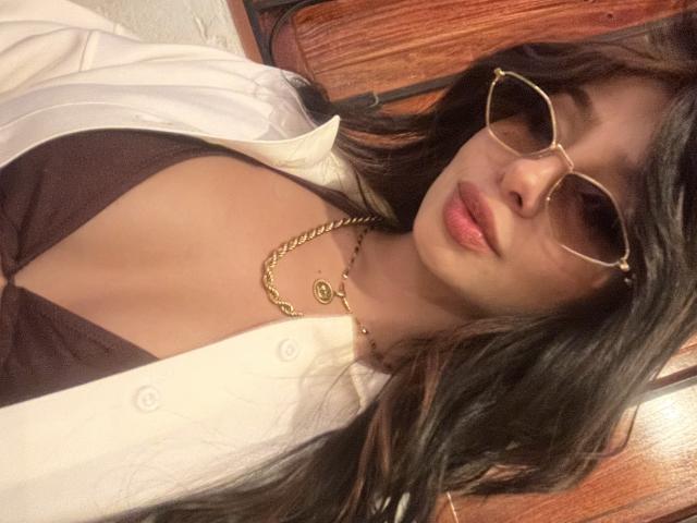 ThaliaCollins with long dark hair, gold-rimmed glasses, white shirt, brown top, gold chain, pouty pink lips, medium tits, slight cleavage, seated on wooden chair, looking at camera.