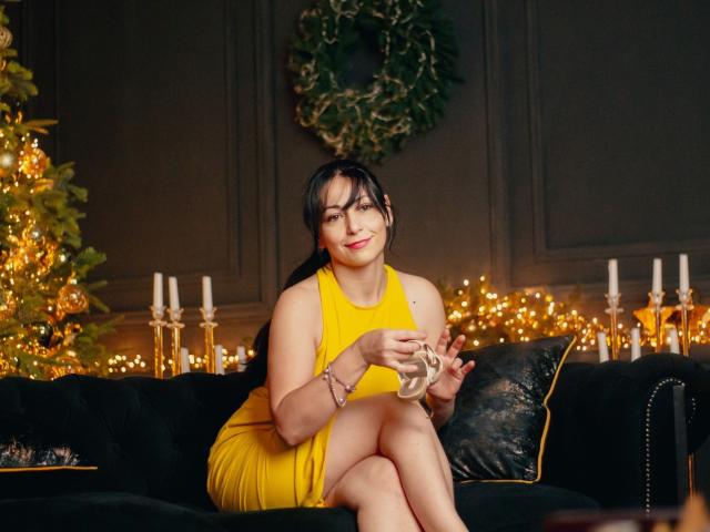 LiluGrants in a yellow dress, sitting on a black couch, holding a glass, smiling, with Christmas lights and candles in the background, showing her ass and tits.