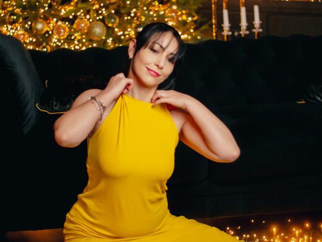 LiluGrants in a bright yellow dress, sitting on a black sofa, smiling, hands near collar, short black hair, pink lipstick, bracelet, Christmas tree with lights in background.