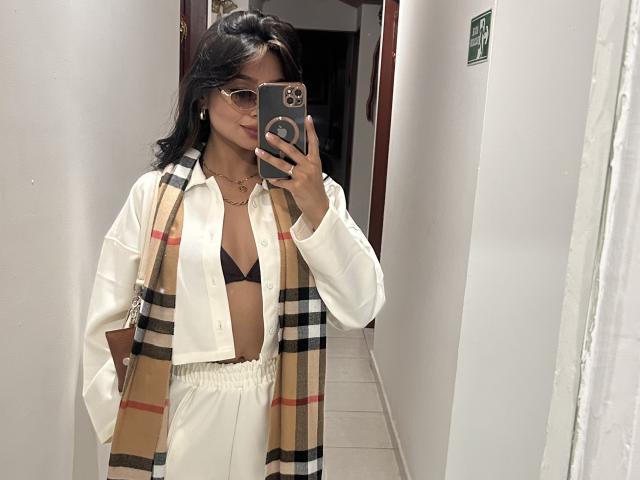 ThaliaCollins with glasses, black bra, white shirt, plaid scarf, white pants, holding phone in mirror, showing medium tits, medium ass, dark hair, standing in hallway.