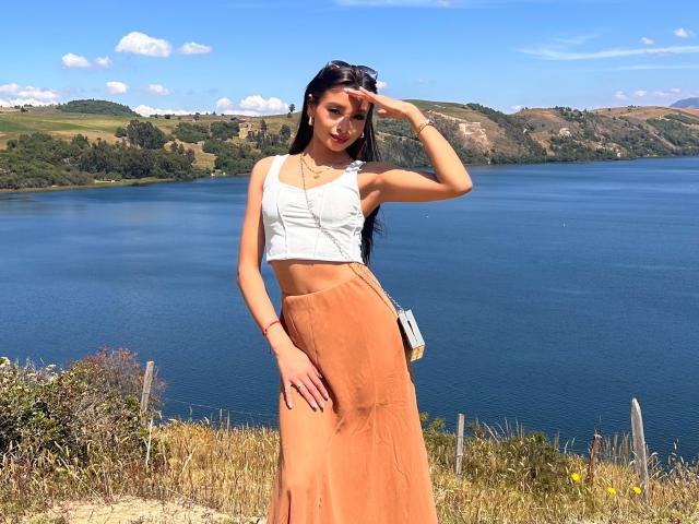 ThaliaCollins with long black hair, wearing a white crop top and orange skirt, standing by a blue lake, hand on forehead, sunglasses on head, smiling, sunny day.