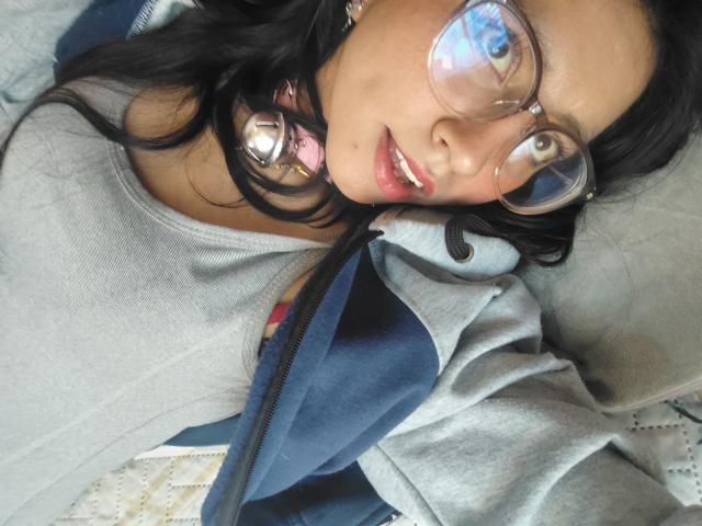 VioletXBloom with glasses, black hair, pink headphones, grey shirt, blue jacket, lying down, looking up, slightly open mouth, soft light, relaxed, casual.