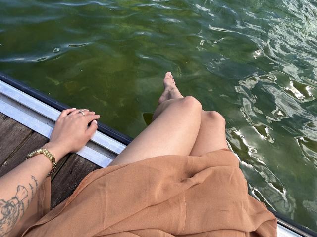 ThaliaCollins' legs in a brown skirt, feet in green water, hands on wooden dock, tattoo on left forearm, gold bracelet, water reflections.