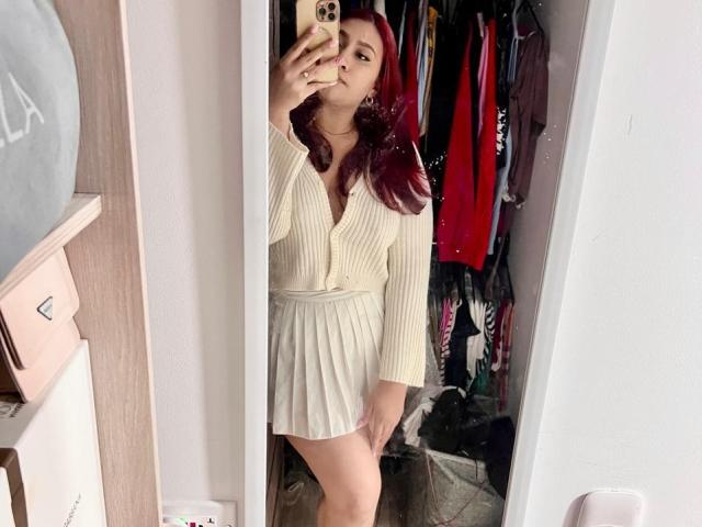 ElyRoss with red hair, wearing a cream ribbed dress, taking a mirror selfie, showing her tits and ass, holding a phone with a gold case.