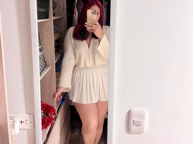 ElyRoss with red hair, wearing a cream ribbed sweater and pleated skirt, taking a mirror selfie, showing her bare thighs and slightly visible ass.