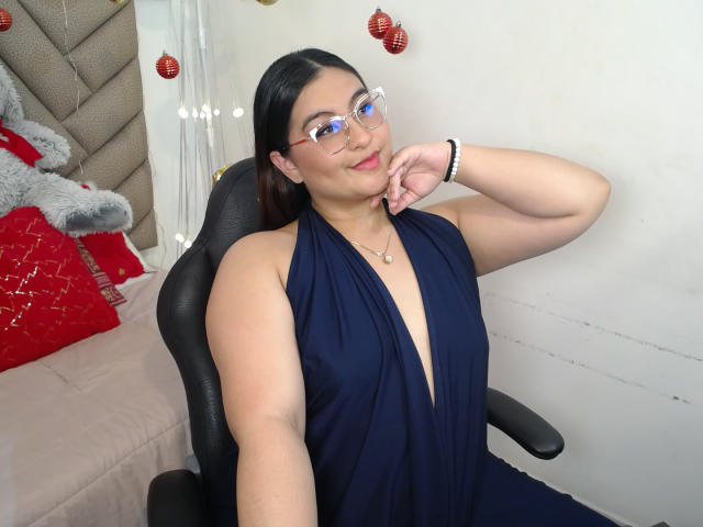 HelueJamil with black hair, glasses, and a deep navy blue halter dress, showing a small cleavage, resting her chin on her hand, sitting in a black chair.