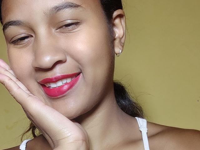 Close-up photo of LoderFist, a woman with medium brown skin, dark hair in a ponytail, and small gold hoop earrings. She's smiling with bright red lipstick, eyes half-closed, and right hand touching her cheek. She wears a white spaghetti strap top. The background is a plain, yellowish wall. LoderFist's face is slightly tilted to the right, and her skin has a smooth texture with subtle natural shine. The image focuses on her facial expression and makeup.