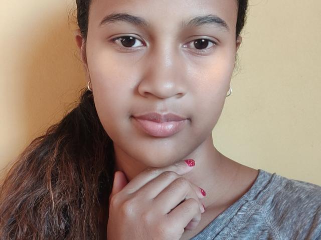 Close-up of LoderFist, a young woman with light brown skin and dark brown eyes. She has long, wavy hair pulled back and is wearing a grey, v-neck shirt. Her right hand is near her chin, with fingers slightly curled and nails painted red with white patterns. She has small, gold hoop earrings. Her lips are slightly parted and she has a subtle, neutral expression. The background is a plain, yellow wall. The lighting is even, highlighting her natural skin texture.