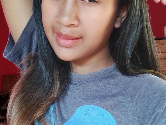 Close-up photo of LoderFist with medium brown skin and long, straight black hair. LoderFist's face is slightly tilted, lips parted with a subtle smile. They wear a gray t-shirt with a blue graphic on the chest. LoderFist's right arm is raised, hand out of frame. Bright red background, with a brown cushioned surface in the lower right. Natural light highlights LoderFist's smooth skin and soft facial features. LoderFist's eyes are slightly open, looking softly at the camera.